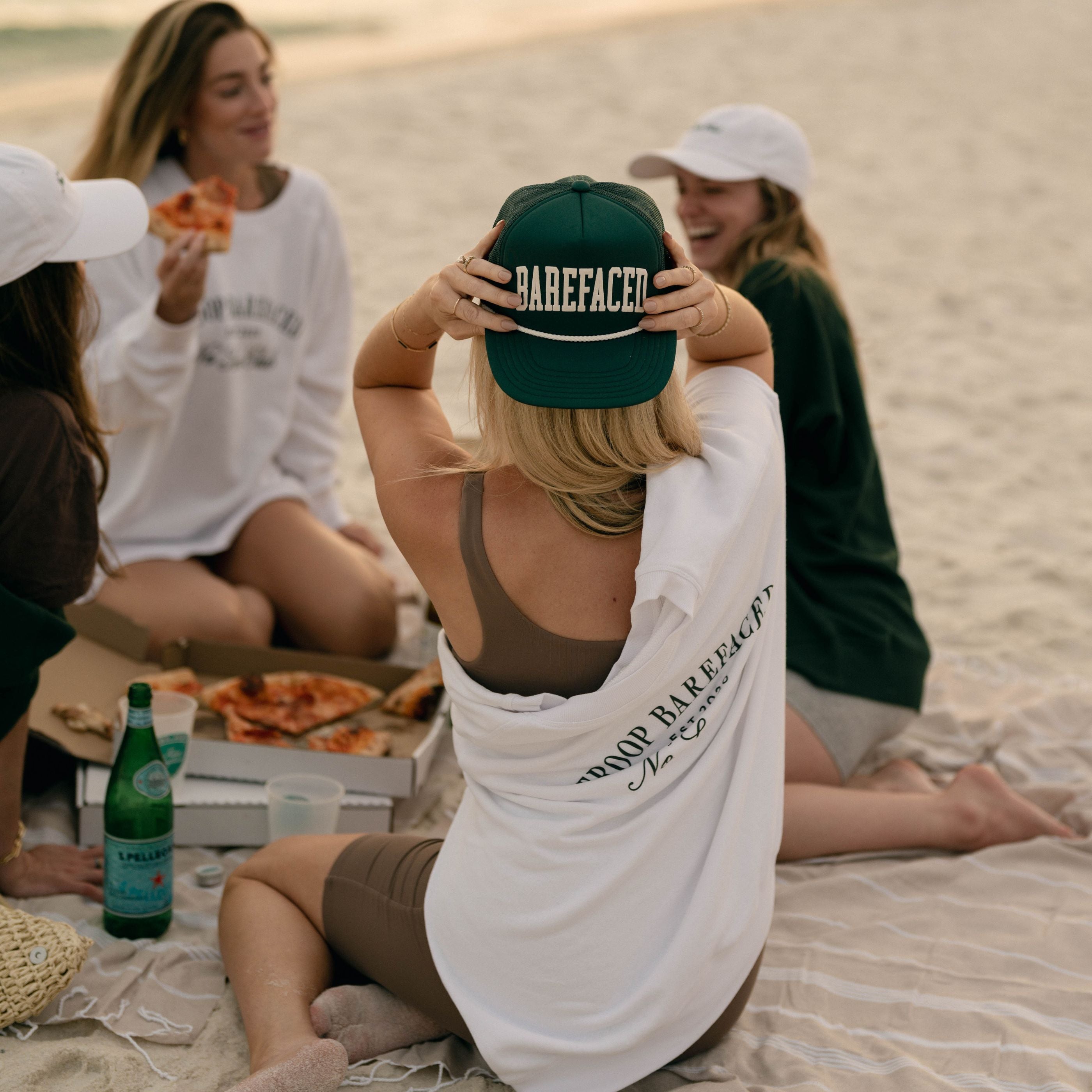 Woman sitting on beach with friends wearing Barefaced Varsity Trucker Hat backwards